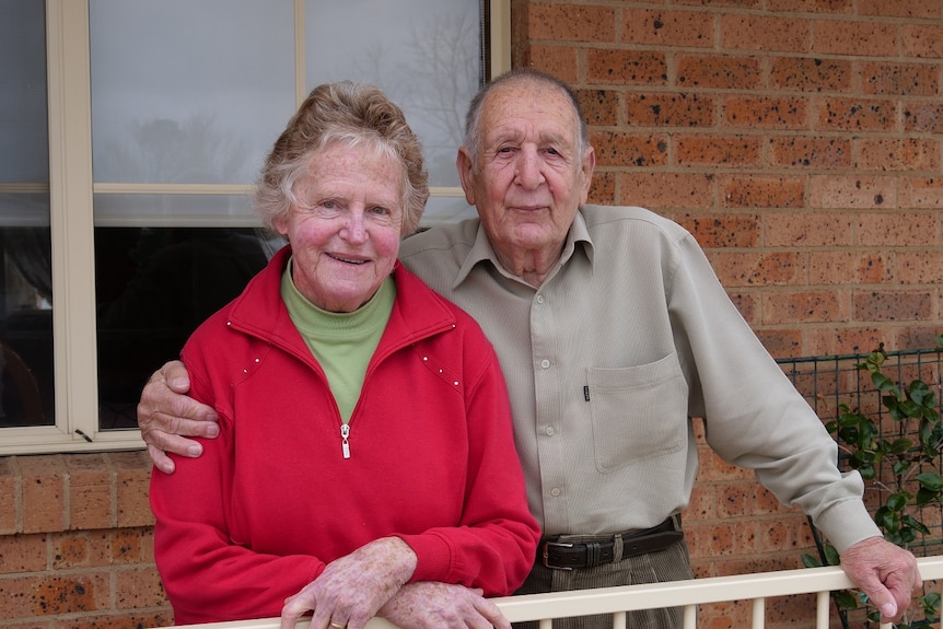 An older couple smile at the camera with their arms around one another from the front balcony of a brick residence