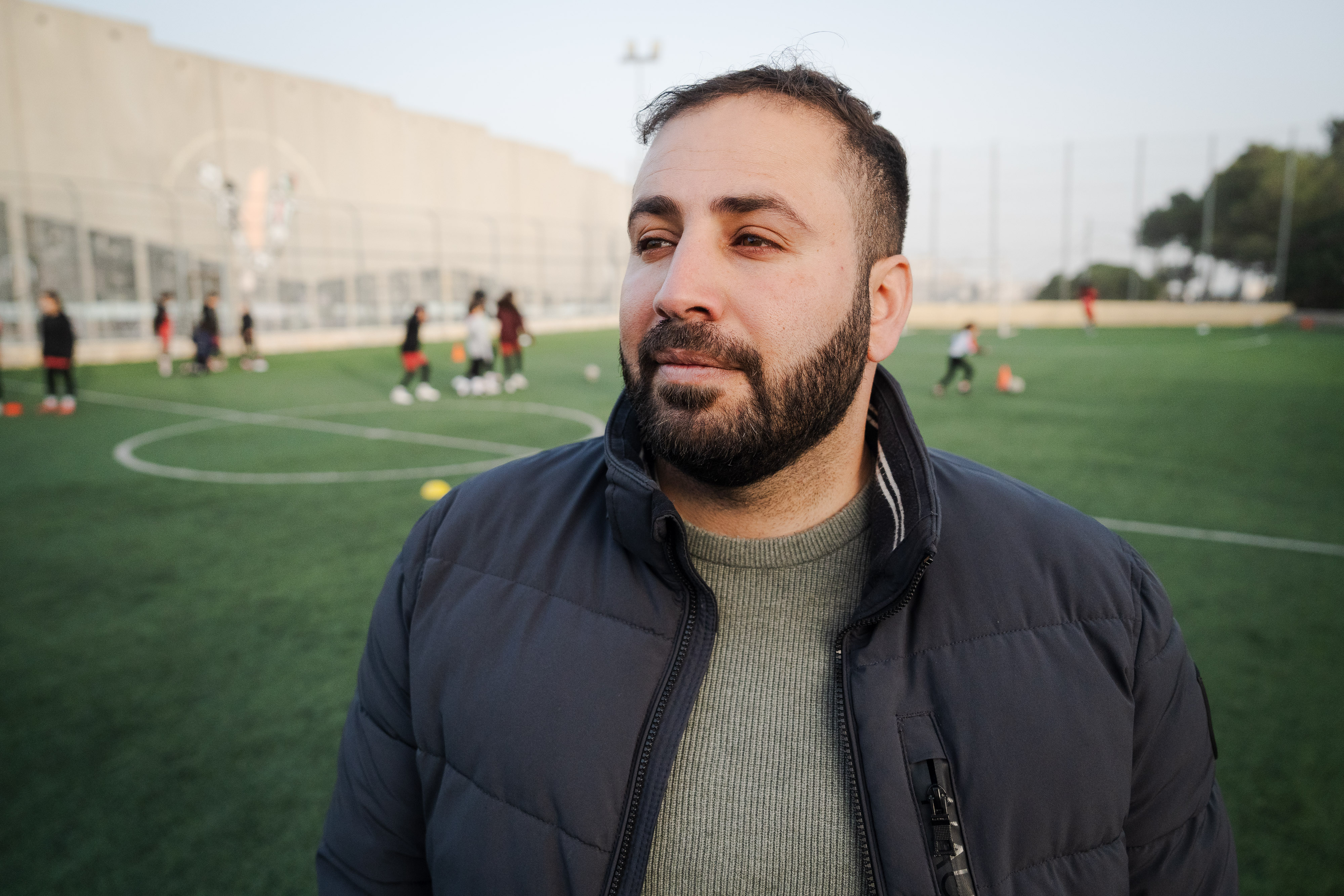 A close-up of a man wearing a dark jacket standing on a soccer field.