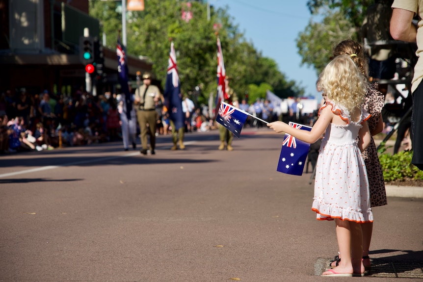 Two young girls holding Australian flags standing along a street, watching troops walk past
