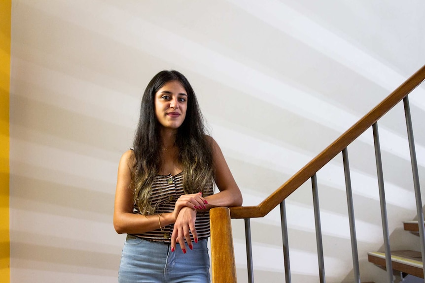 A woman in a striped singlet and jeans leans against a staircase banister in front of a white wall with a yellow stripe.