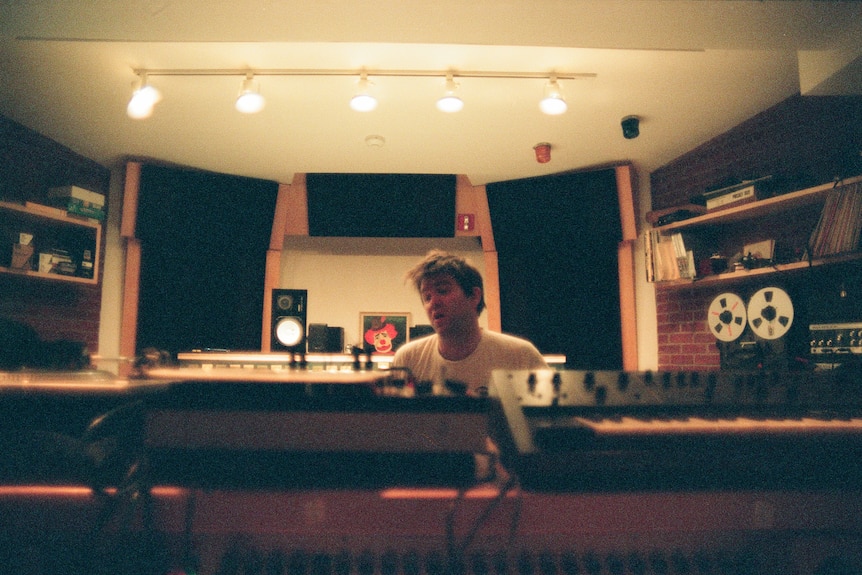 A shaggy-haired man in his 30s, wearing a white T-shirt, is visible over the top of musical equipment, including a keyboard