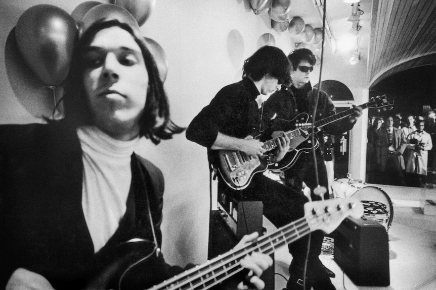 A black and white image of The Velvet Underground on stage, with John Cale on bass, and Sterling Morrison and Lou Reed on guitar