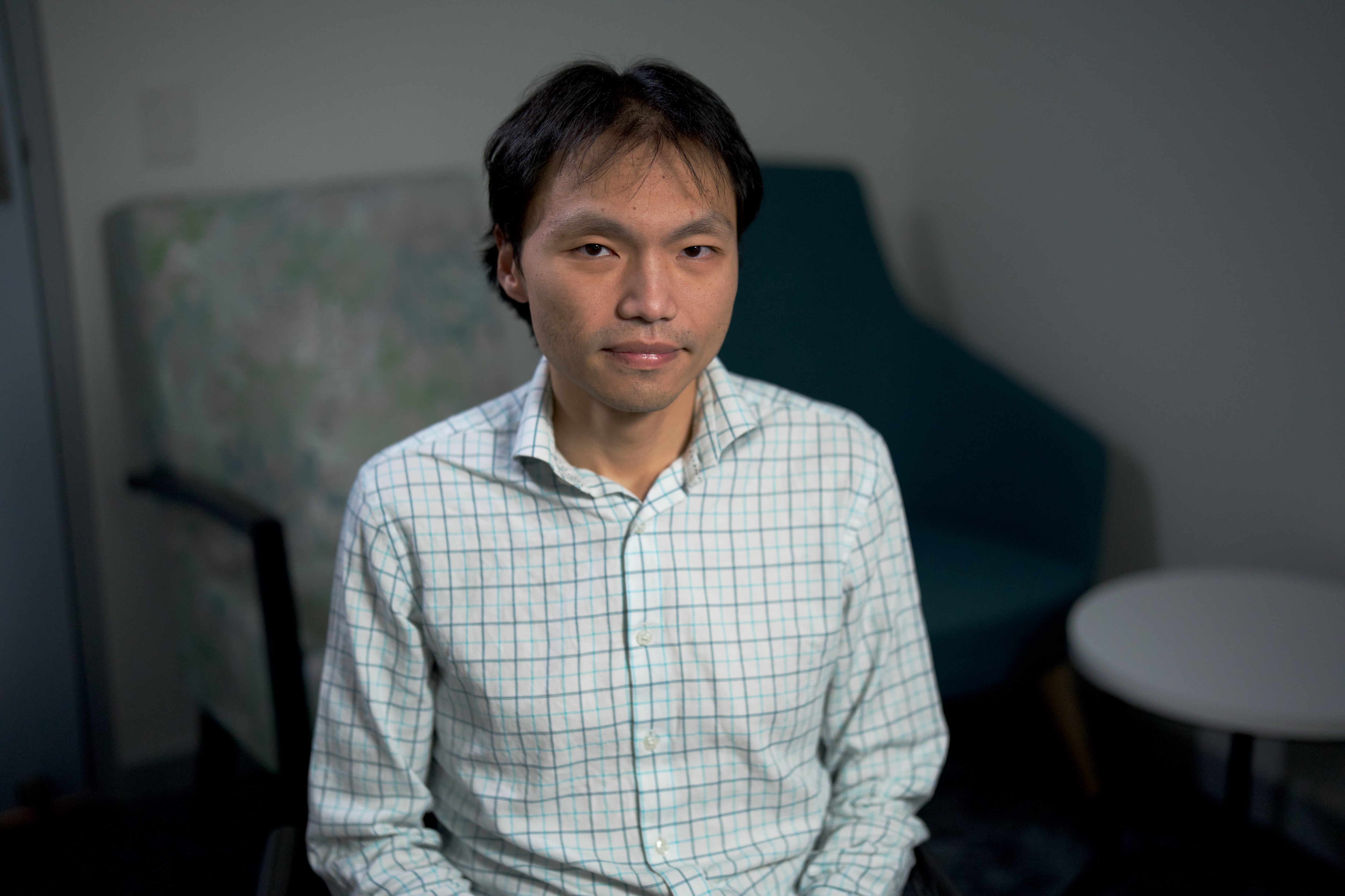 An Asian man in a white shirt with a checked pattern sitting in a chair.