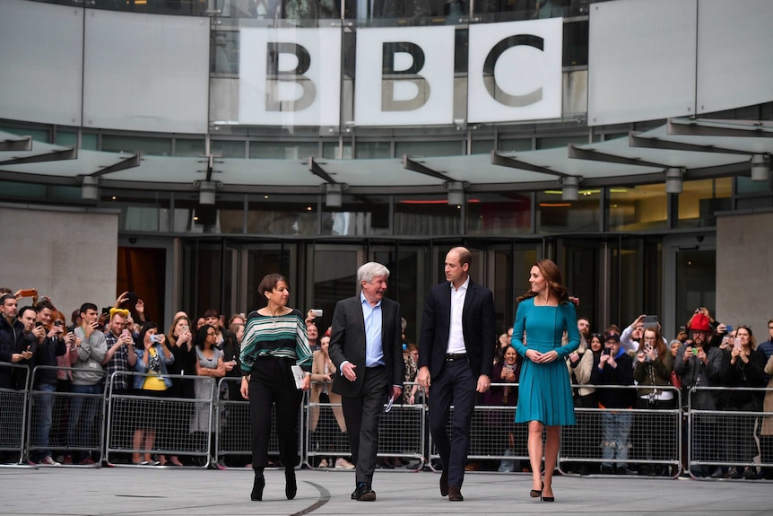You view Prince William and Kate, Duchess of Cambridge, walk with BBC staff in front of crowds under a large BBC sign.