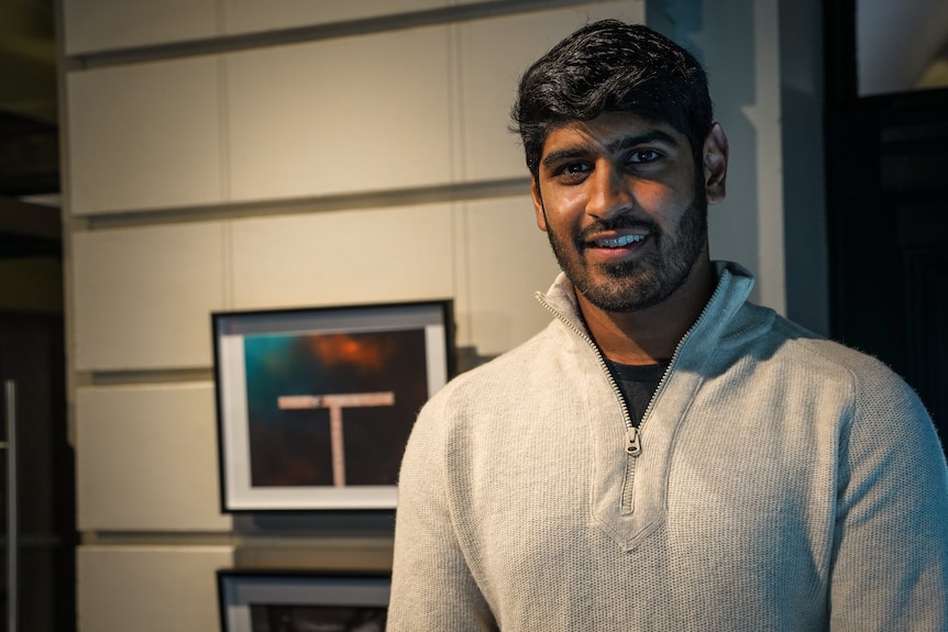 Prashanth Naidoo smiles as he stands, in a jumper in a gallery beside his photography art.