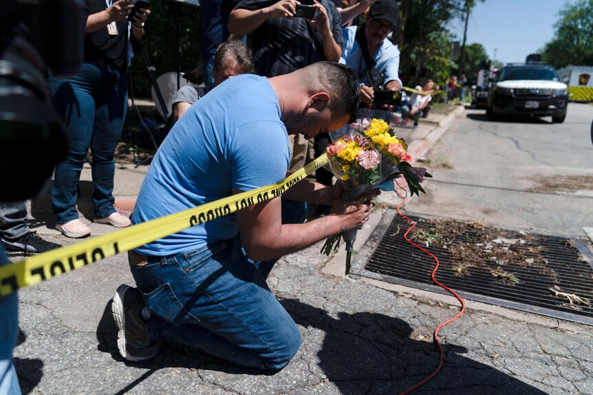 A man kneels next to a yellow police tape holding a bouquet of flowers with his head bowed