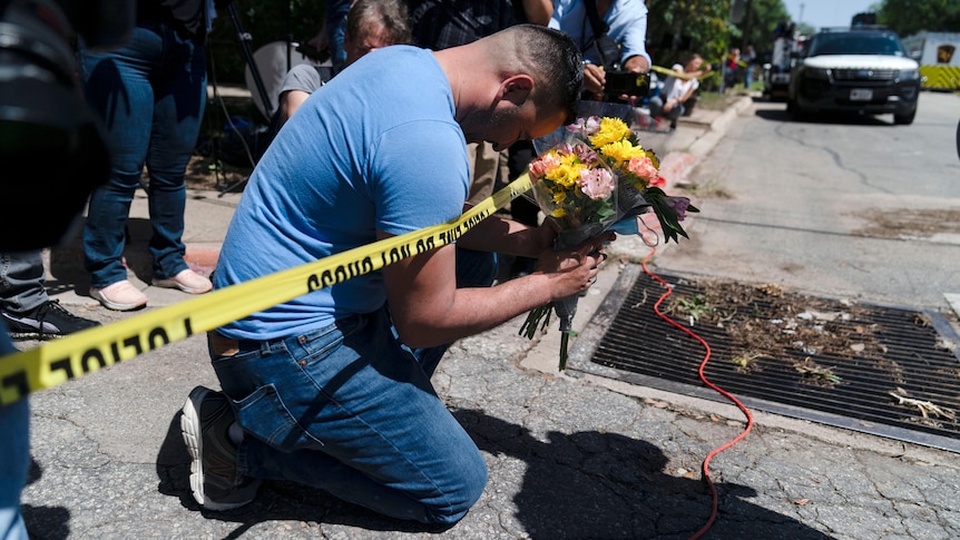 A man kneels next to yellow police tape holding a bunch of flowers with his head bowed