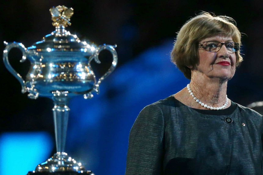 Margaret Court, wearing a formal dress and string of pearls, stands beside the Australian Open trophy at a victory ceremony.