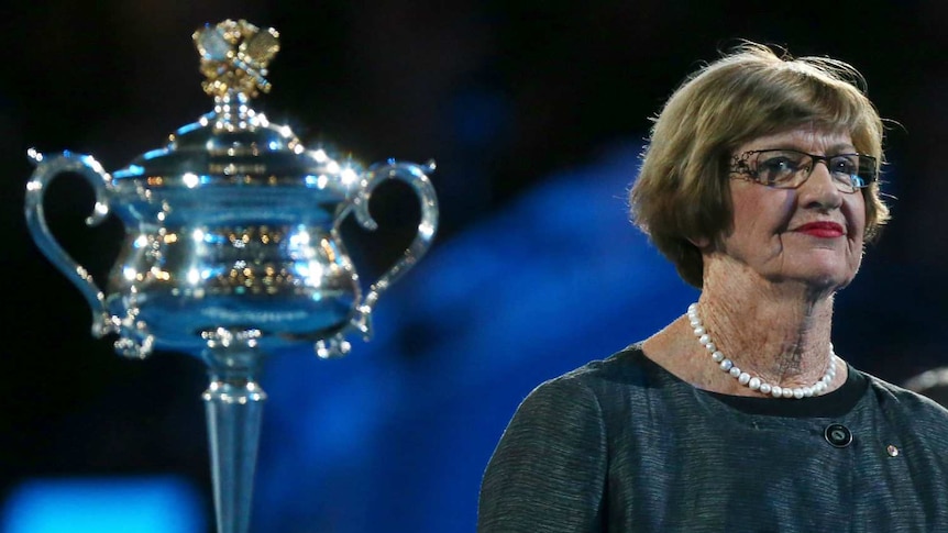 Margaret Court, wearing a formal dress and string of pearls, stands beside the Australian Open trophy at a victory ceremony.