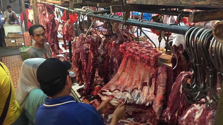 Three people standing at a market stall in Indoneisa, with meat hanging from hooks.