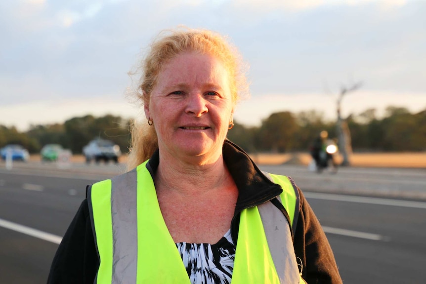 A serious middle-aged woman with blond hair tied back in a high viz vest in front of a road.
