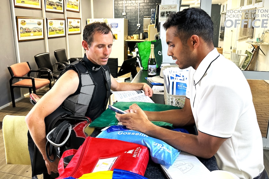 A man with brown skin wearing a white shirt looking at his mobile phone talks to a man wearing a jockey vest holding a saddle.