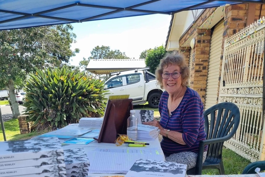 A smiling, bespectacled woman with curly hair sits outside at a table upon which copies of a book are stacked. 