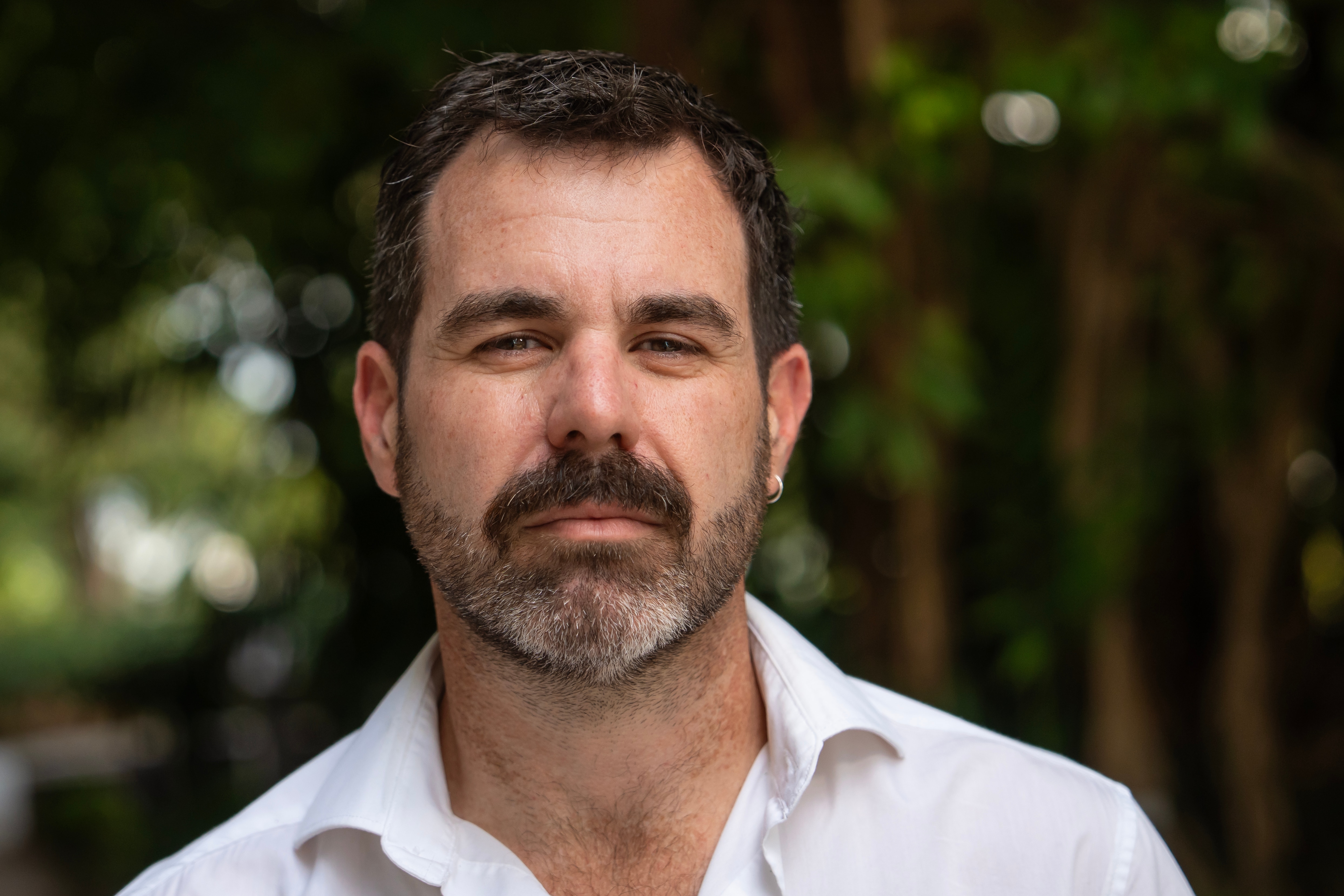 A man with dark brown hair, eyes and beard, moustache, wearing a white collared shirt, serious expression on his face.