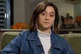 A young woman with short brown hair sits on a couch, wearing a white turtleneck and denim jacket.