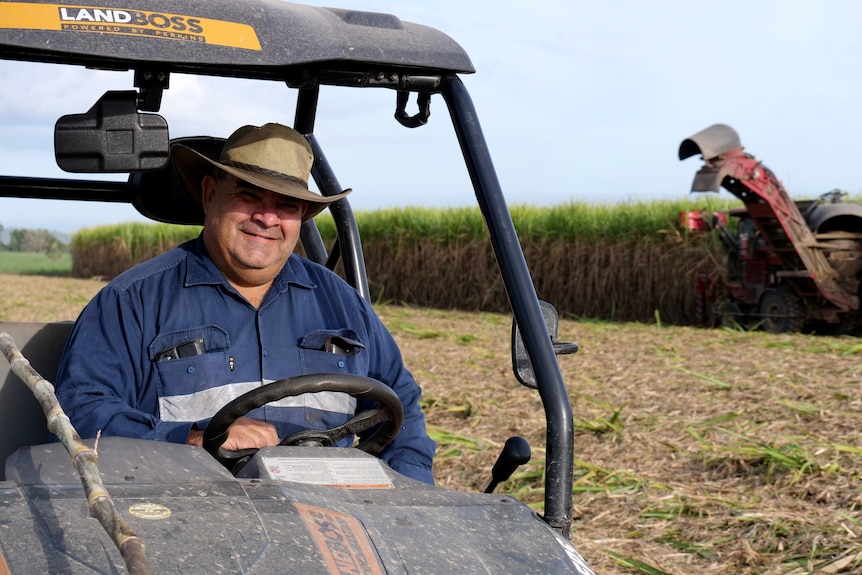 A man in a broad rimmed hat sits on an ATV, with sugar cane being harvested in the background. 