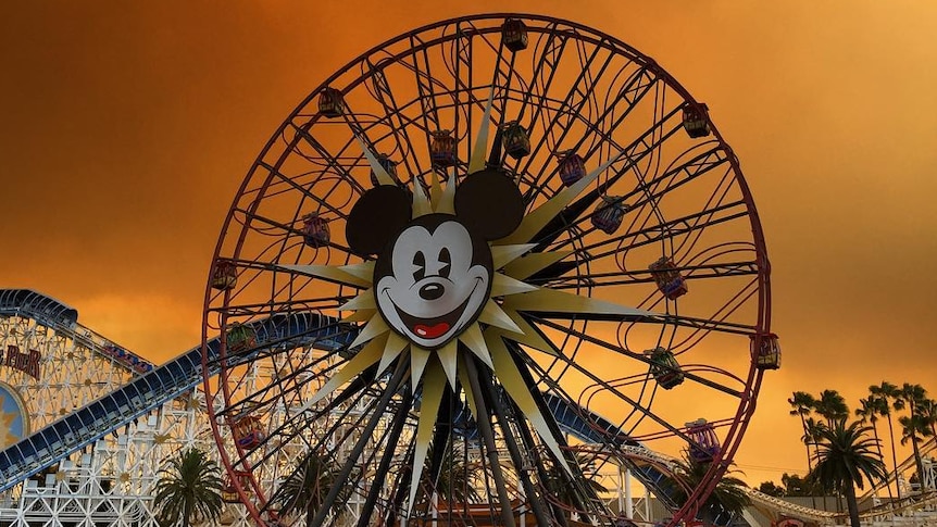 An orange sky with dark smoke is seen over the rollercoasters and Mickey Mouse ferris wheel at Disneyland.