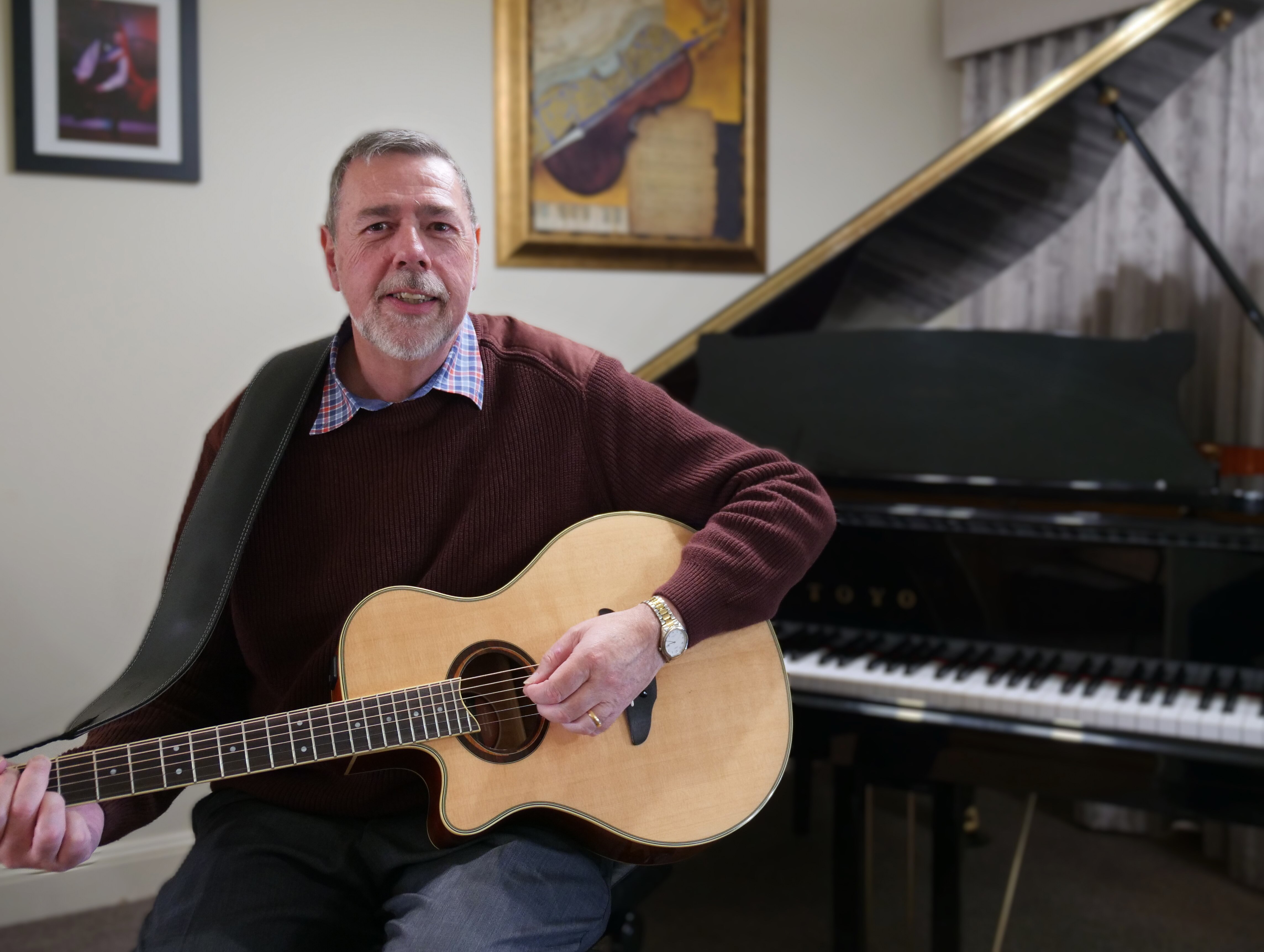 A salt and pepper haired man sits in front of a grand piano, cradling a guitar and smiling.