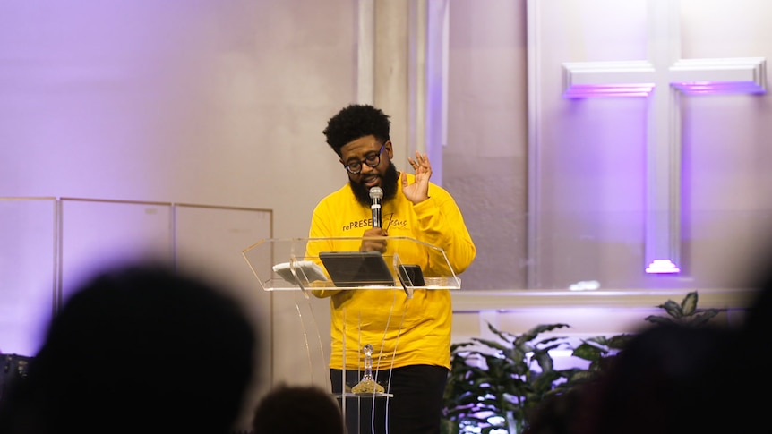 A Black man wearing a yellow shirt delivers a speech in front of a podium on a stage.