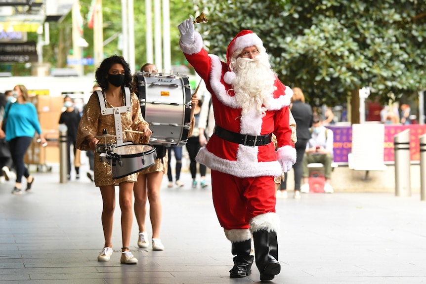Santa Claus walks down a mall, ringing a bell. A woman waring a facemask plays drums behind him.
