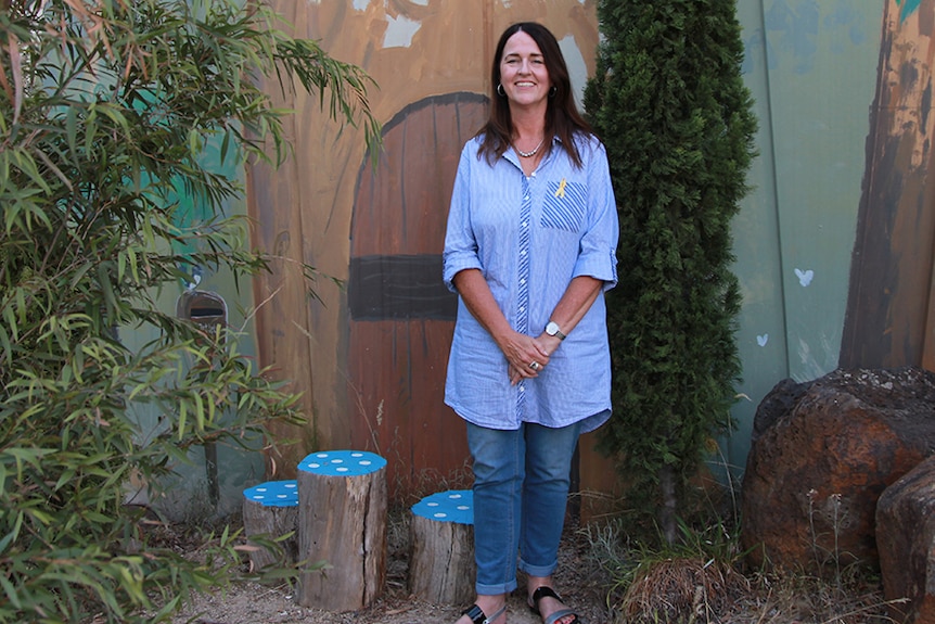 A woman in a blue shirt and jeans stands in a garden in front of a mural of a fairy house.