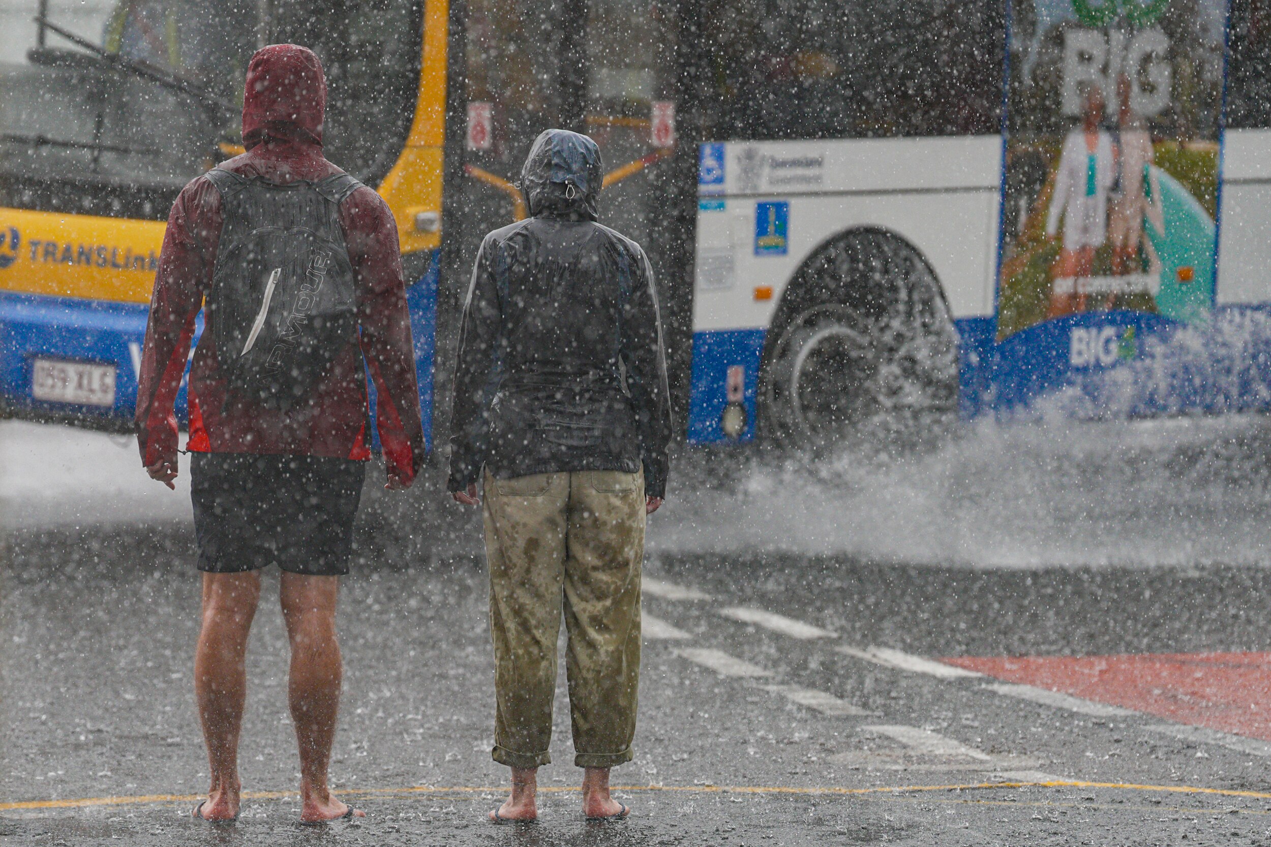 Two people wearing rain jackets waiting for a traffic light in the city, a passing bus splashes a puddle of water.