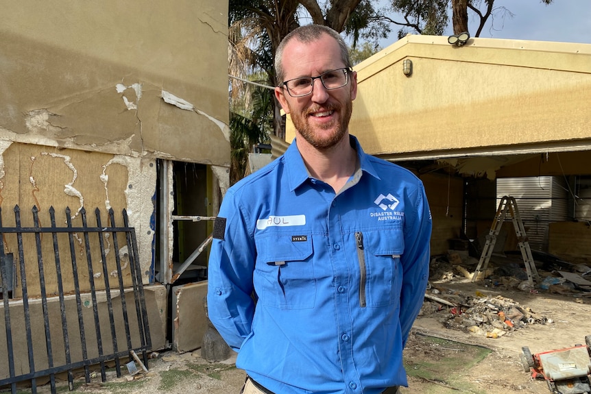 A man has short brown hair and a red beard. he wears glasses and a blue work shirt. He stands in front of  water damaged house