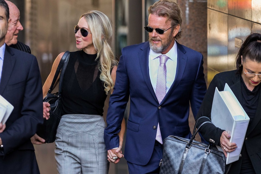 Craig McLachlan, dressed in a suit, arrives at court, flanked by lawyers and holding the hand of his partner.
