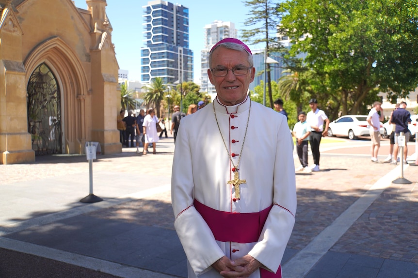 A man in glasses donning a white and red ensemble with a gold cross around his neck smiles to the camera