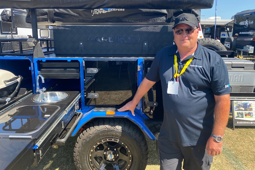 Middle aged man in dark blue polo and black cap standing in front of blue off-road camping trailer
