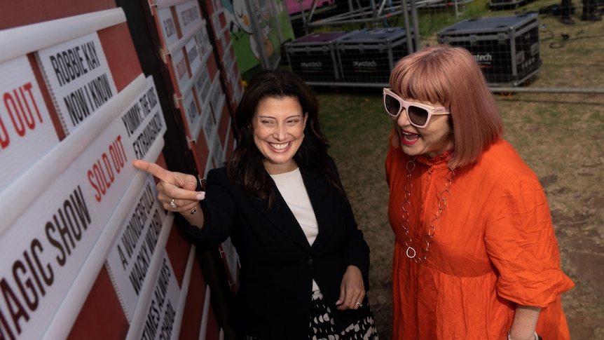 Andrea Michaels points at a 'sold out' card on a board advertising shows with Heather Croall standing next to her smiling