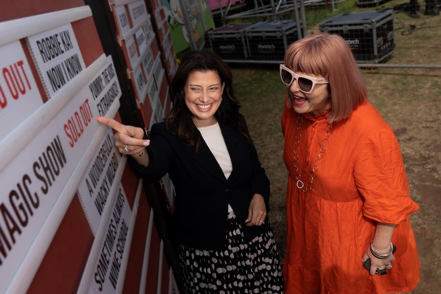Andrea Michaels points at a 'sold out' card on a board advertising shows with Heather Croall standing next to her smiling