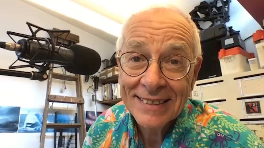 Man in colourful shirt smiling at camera, surrounded by microphone and broadcast equipment.