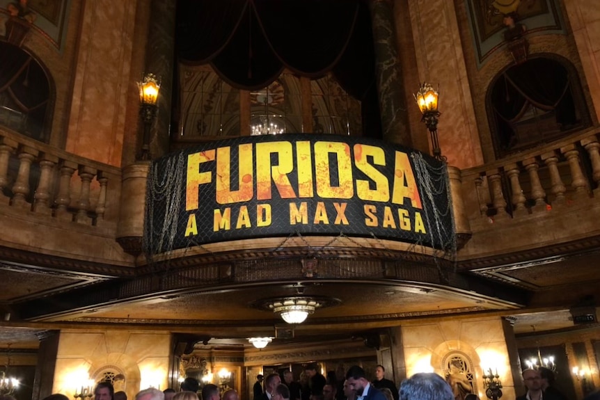 A big banner that reads 'Furiosa, a Mad Max saga' in yellow on a balck background hangs over an ornate balcony.