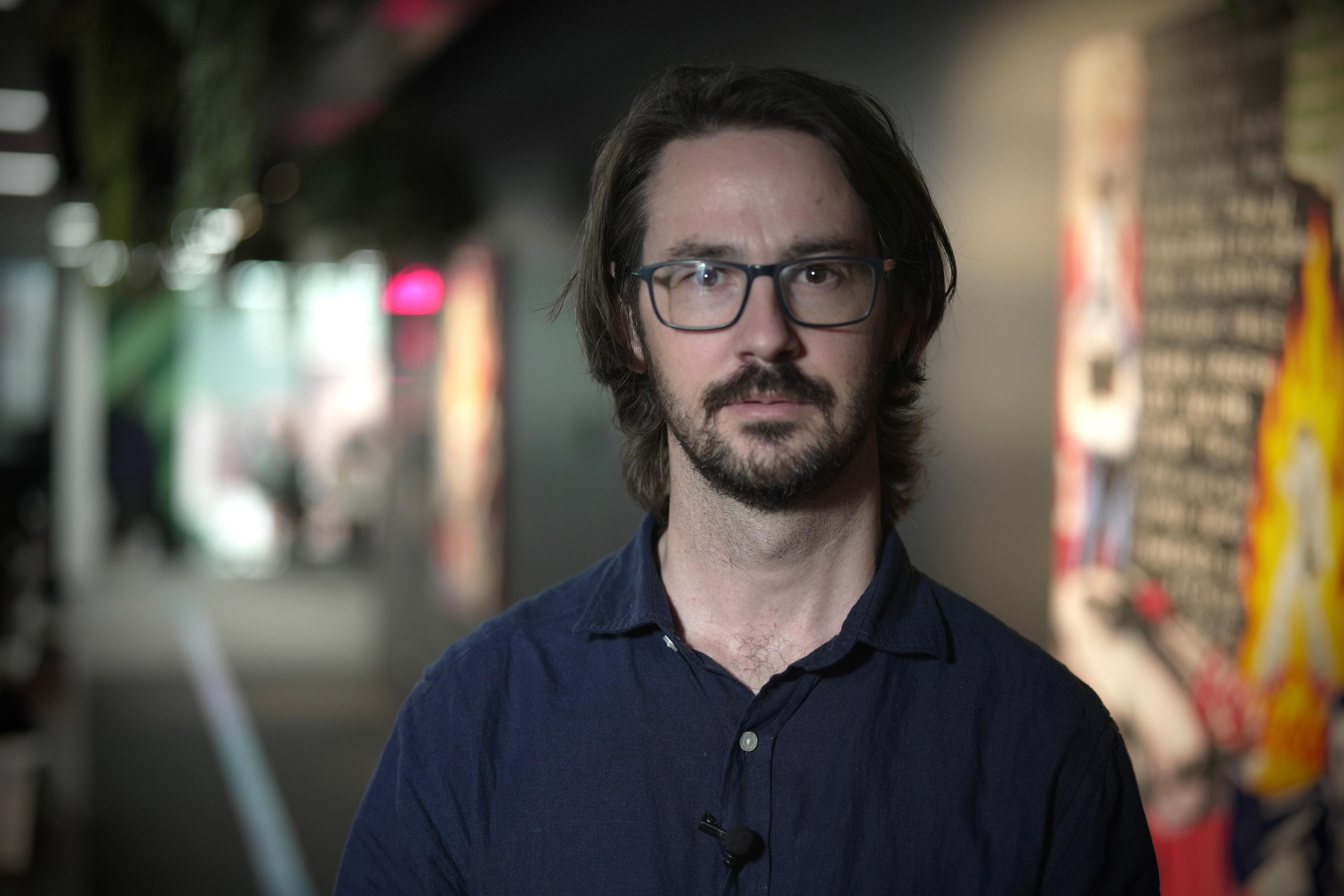 Um homem com camisa de colarinho azul-marinho, barba e óculos está em um corredor de escritório moderno e colorido.