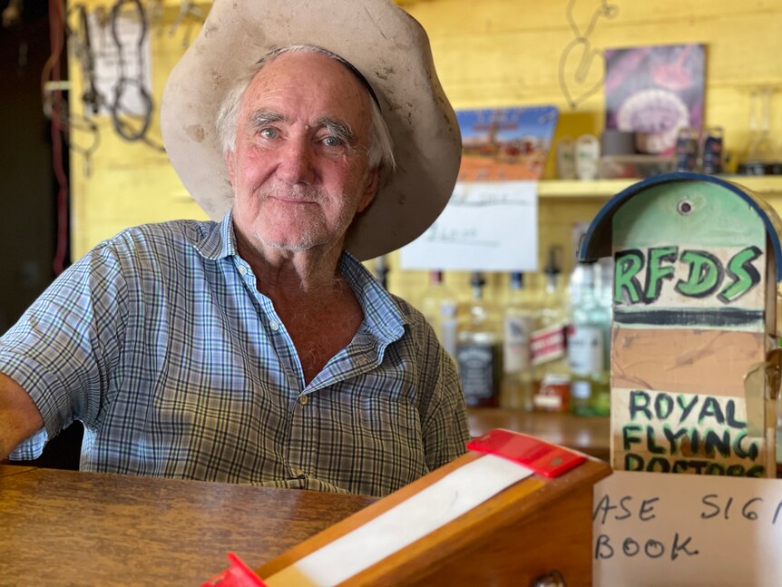 An elderly man with a big white white sits behind a bar, with a sign in book and RFDS fundraiser in the foreground.