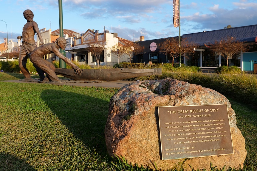 A bronze statue depicting two men pulling a canoe with a rock in front with a description.