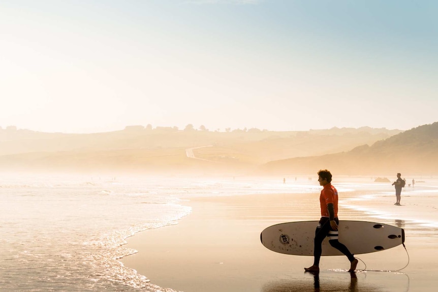 A man walks out to the ocean with a surfboard to depict how to choose your first surfboard.