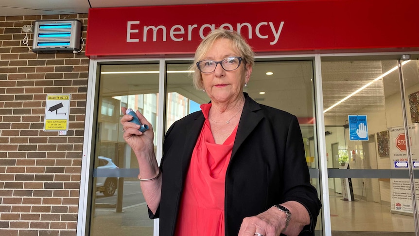 A woman with short hair and wearing glasses, holds an asthma puffer in front of Wagga Wagga Base Hospital emergency department.