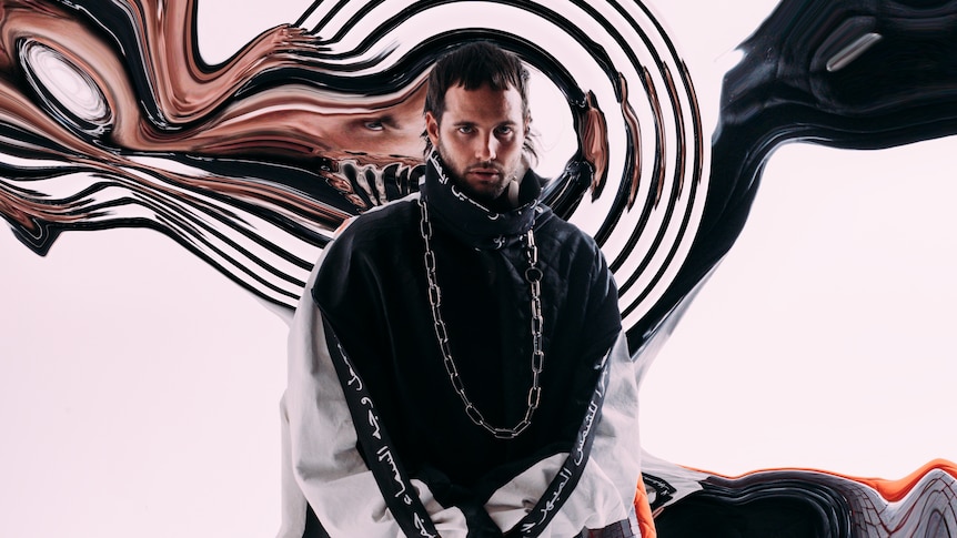 What So Not wears a baggie black windbreaker with white sleeves & stands in front of black & bronze swirls on a white wall.