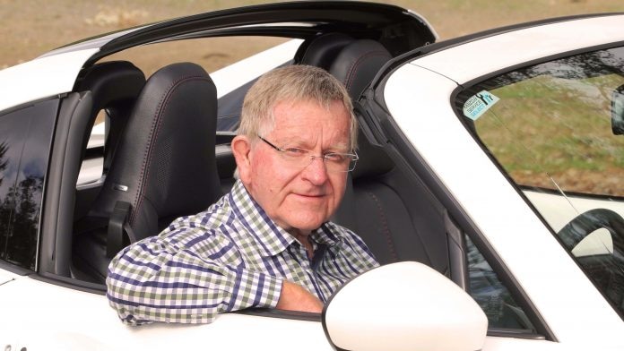 A man with grey and glasses sits behind the wheel of a white convertible
