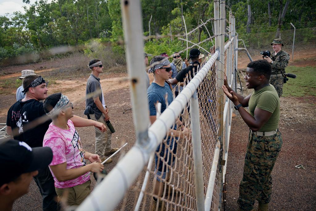 Two groups gathered on opposing sides of a cyclone fence, one in civilian clothes and the other in army uniform