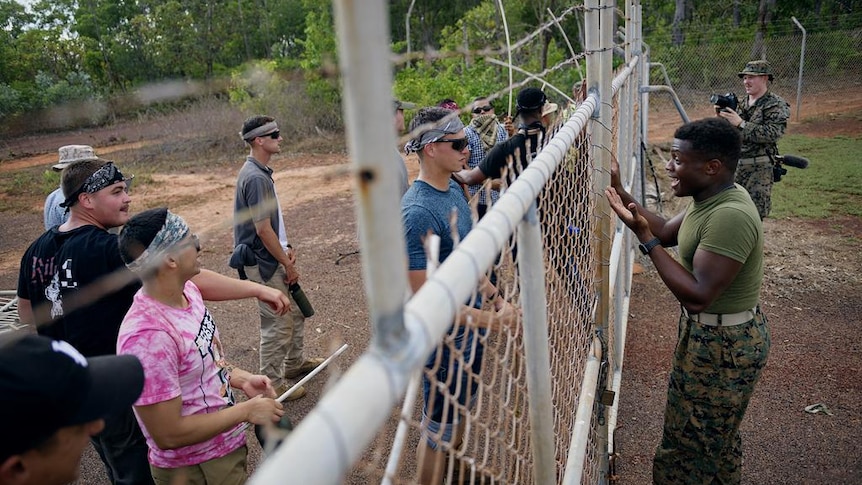 Two groups gathered on opposing sides of a cyclone fence, one in civilian clothes and the other in army uniform