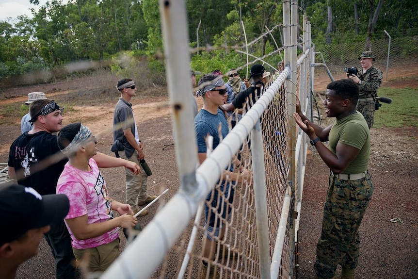 Two groups gathered on opposing sides of a cyclone fence, one in civilian clothes and the other in army uniform