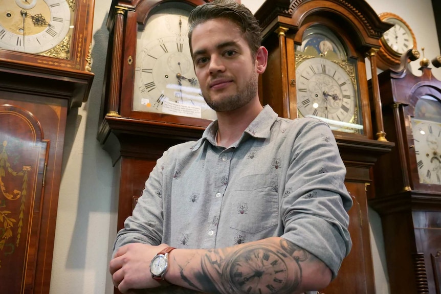 Tim Stewart, who has a large tattoo of a clock on his arm, stands in front of large clocks in a shop