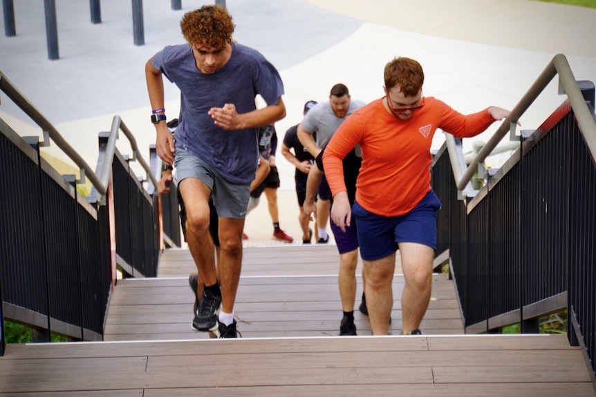 A group of young men in fitness attire run up a flight of stairs outdoors.
