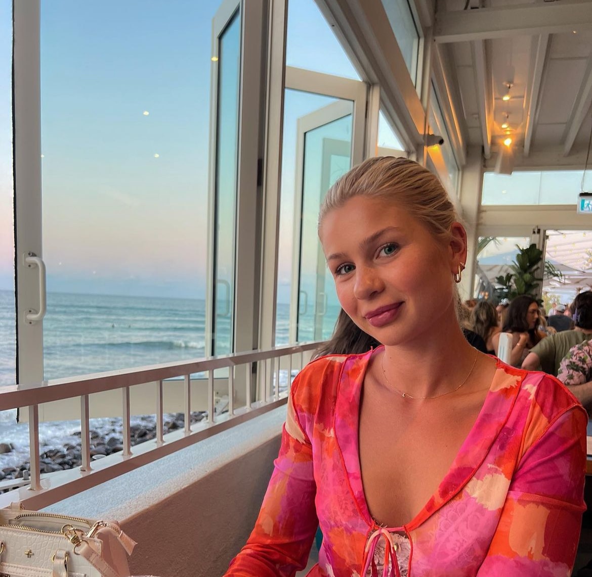 A blonde girl smiles at the camera sitting ihn a bar with the beach in the background. 