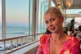 A blonde girl smiles at the camera sitting ihn a bar with the beach in the background. 