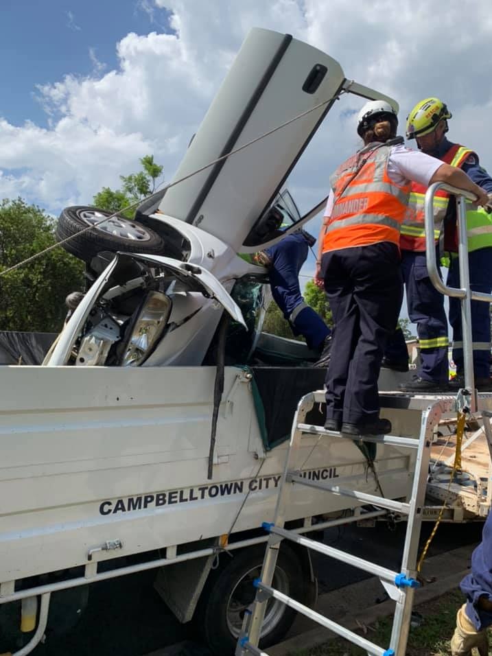 Fire and rescue workers stand on a ladder looking down at the car in the truck tray.