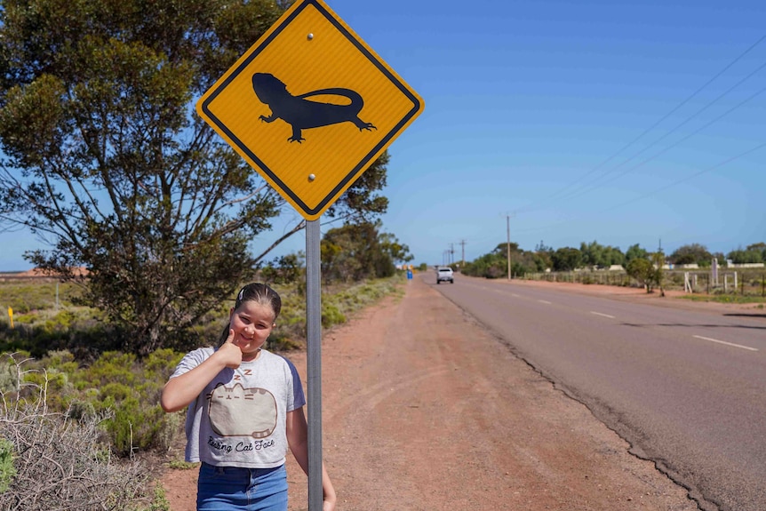 A little girl with brown hair and her thumb up on the side of the road standing next to a lizard sign. 
